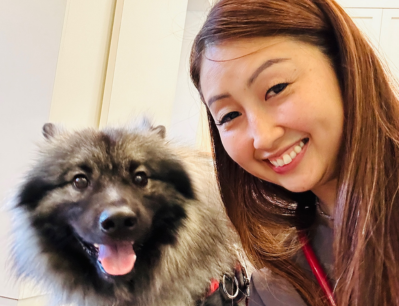 A woman in a black shirt sits on a couch, accompanied by a dog.